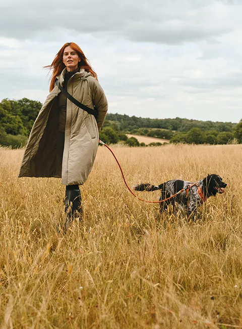 Vrouw wandelt met haar hond door een veld, gekleed in lange jas van Sweaty Betty.