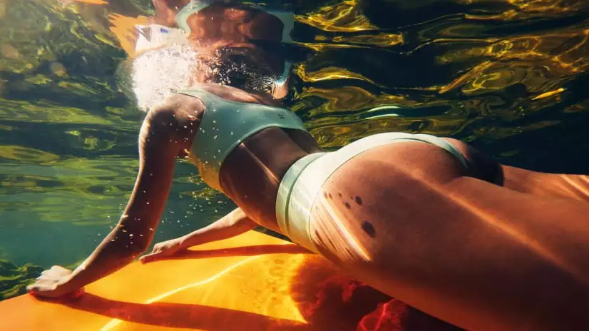 Onderwaterfoto van vrouw leunend op een oranje surfboard in helder water.