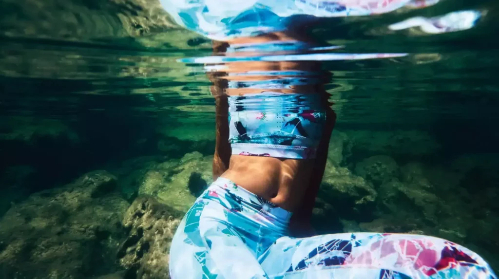 Onderwaterfoto van vrouw in kleurrijke sportlegging tussen rotsen.
